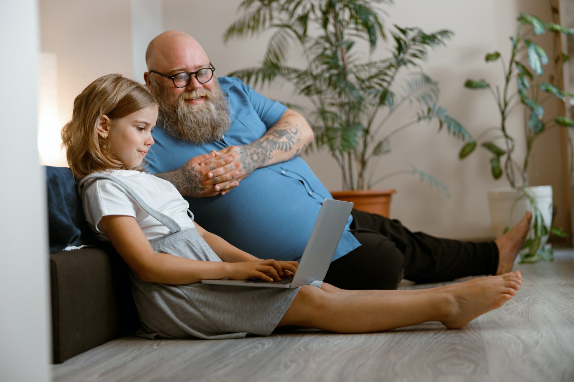 Smiling dad and cute girl watch video via laptop sitting on floor in bedroom